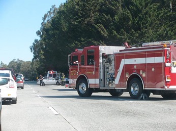 View from the perspective of an approaching motorist of a highway accident scene where a firetruck blocks the right lane and traffic flows in the left lane